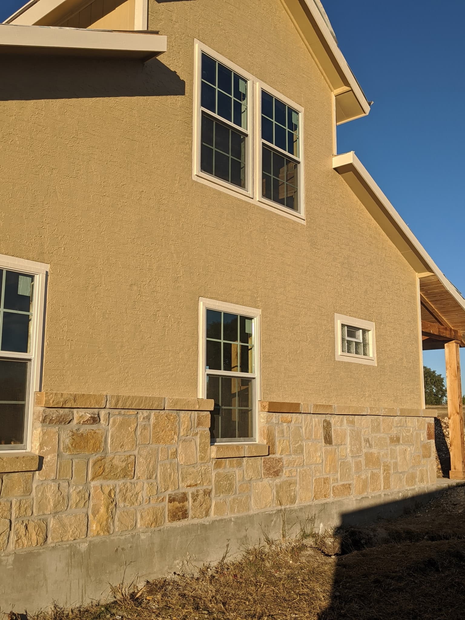 Tan stucco house with light stone veneer base and white-trimmed windows under a clear sky.