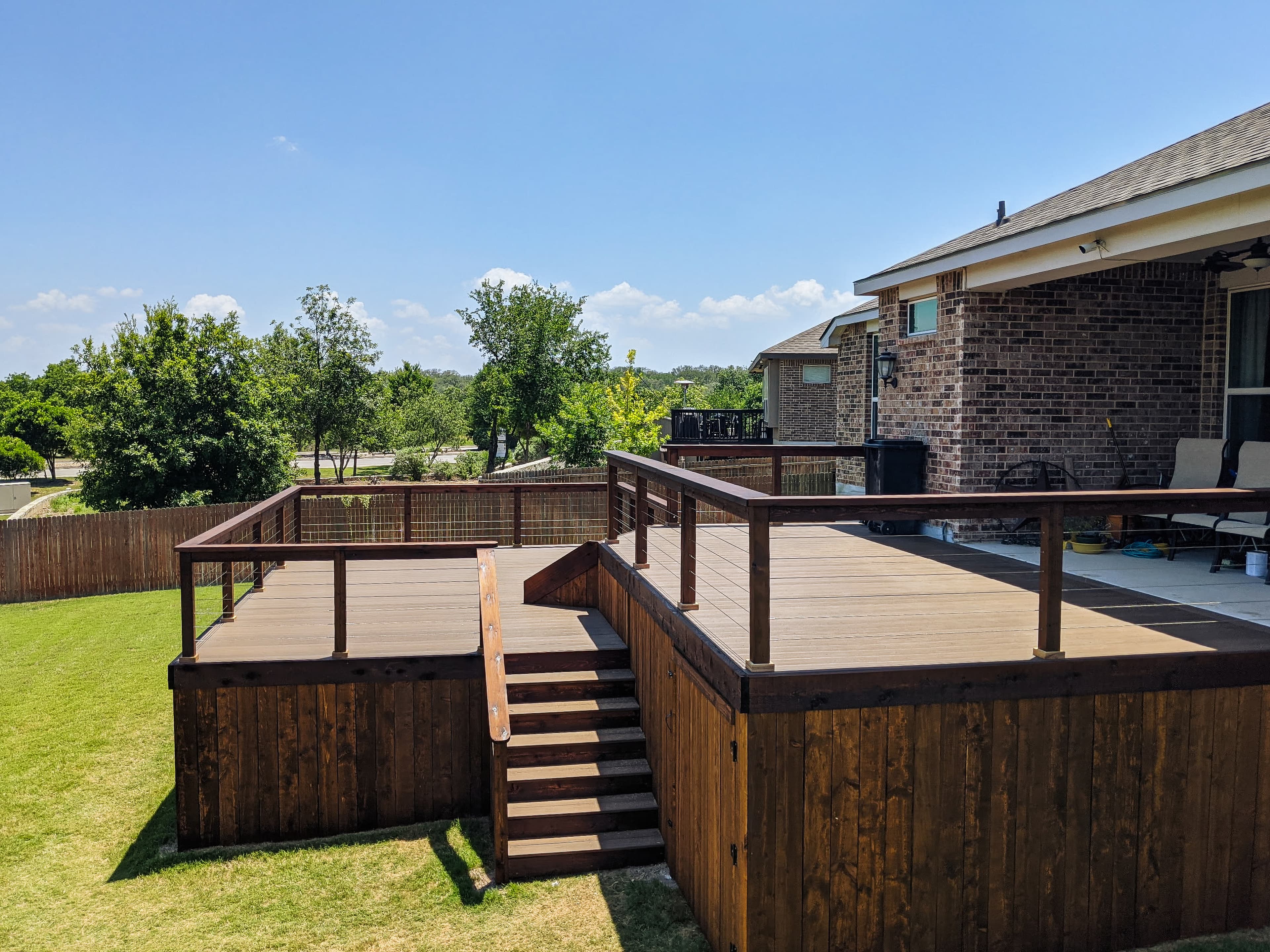 Multi-level dark wood deck with wire railings attached to a brick house in green backyard.