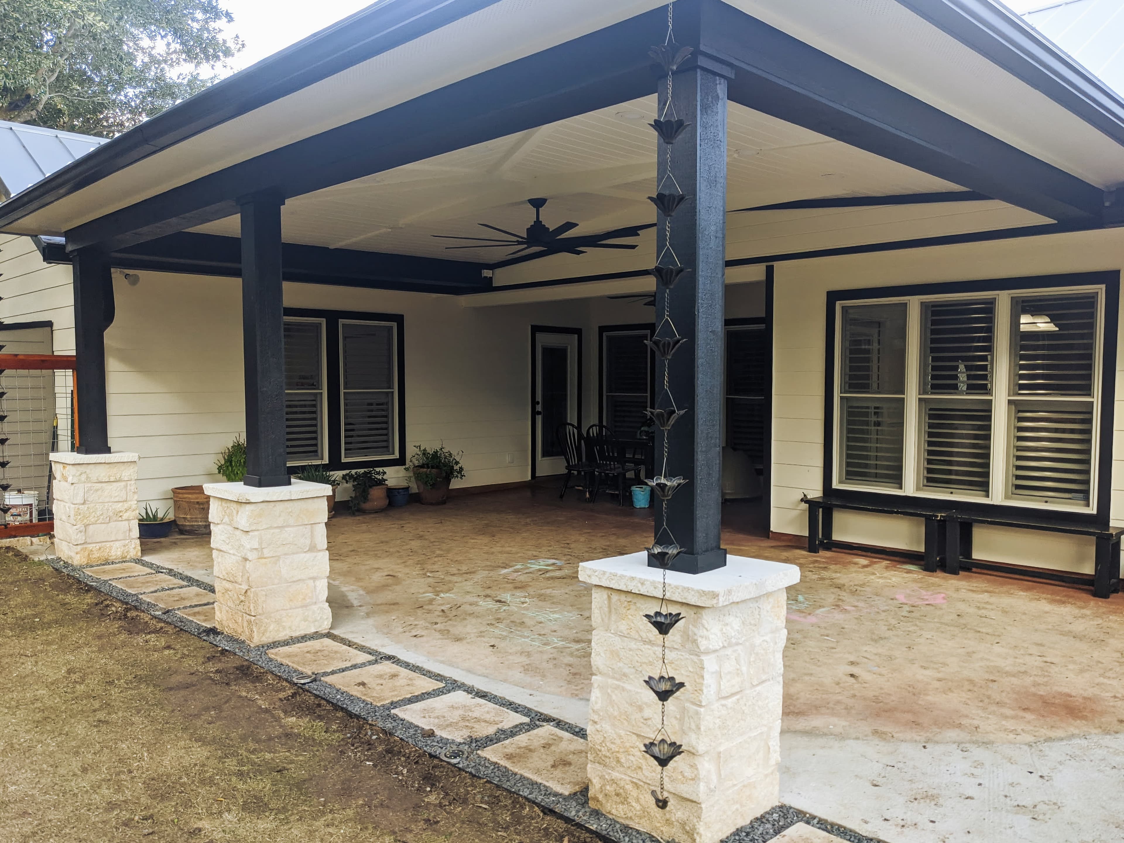 Covered patio with black beams, stone column bases, a ceiling fan, and decorative rain chain.