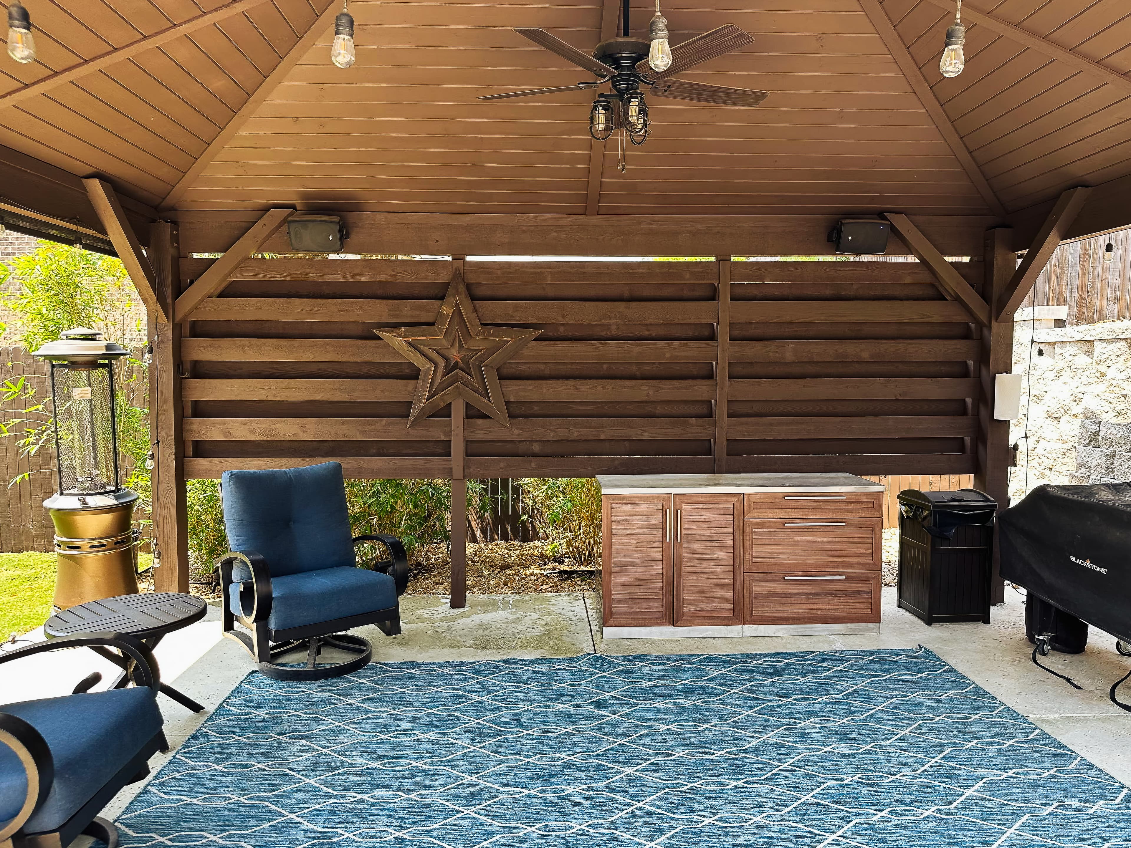Wooden gazebo patio featuring blue armchairs, a patterned rug, outdoor kitchen cabinets, and a grill.