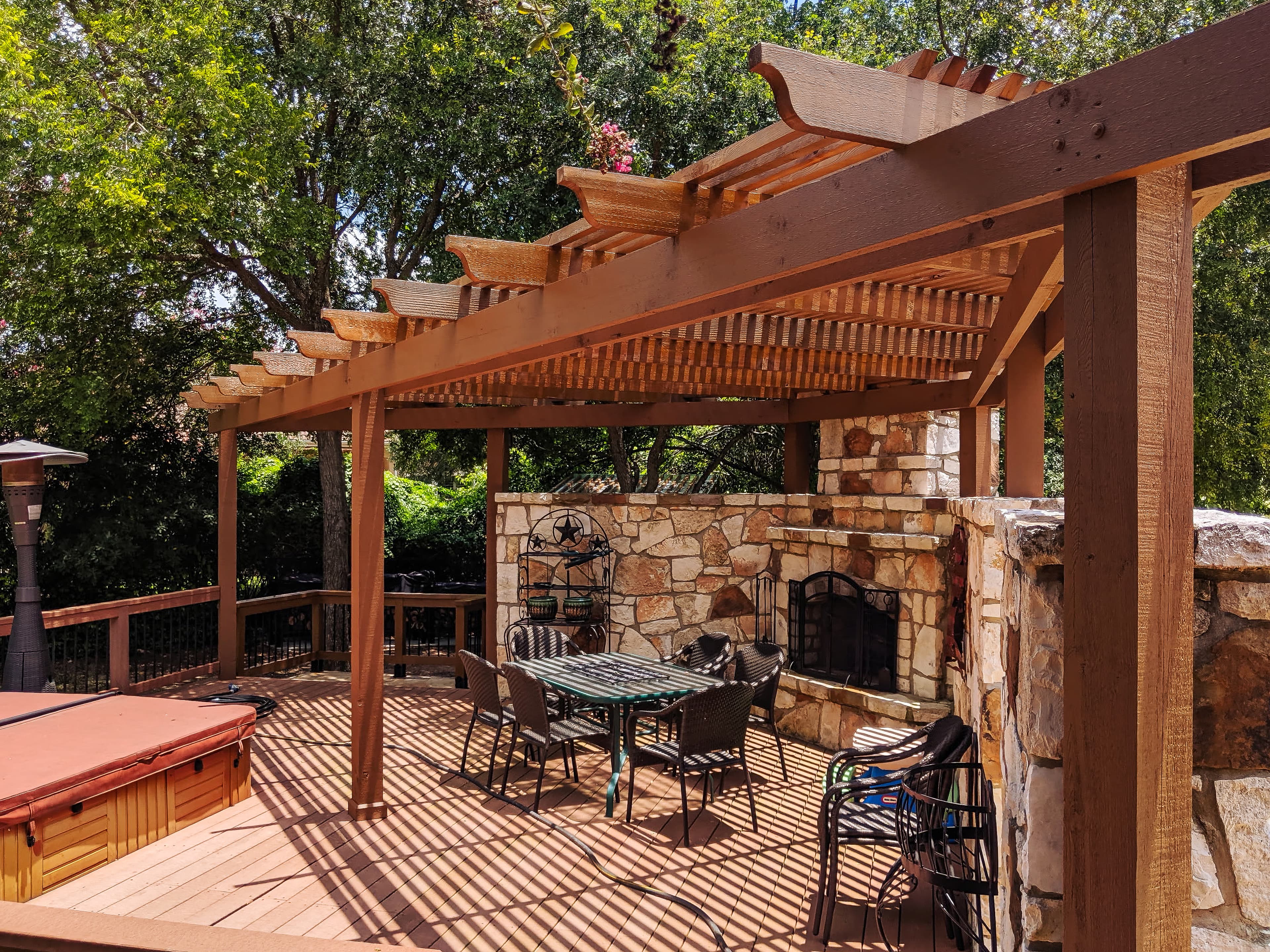 Large wooden pergola shades a stone fireplace and dining area on a backyard deck.