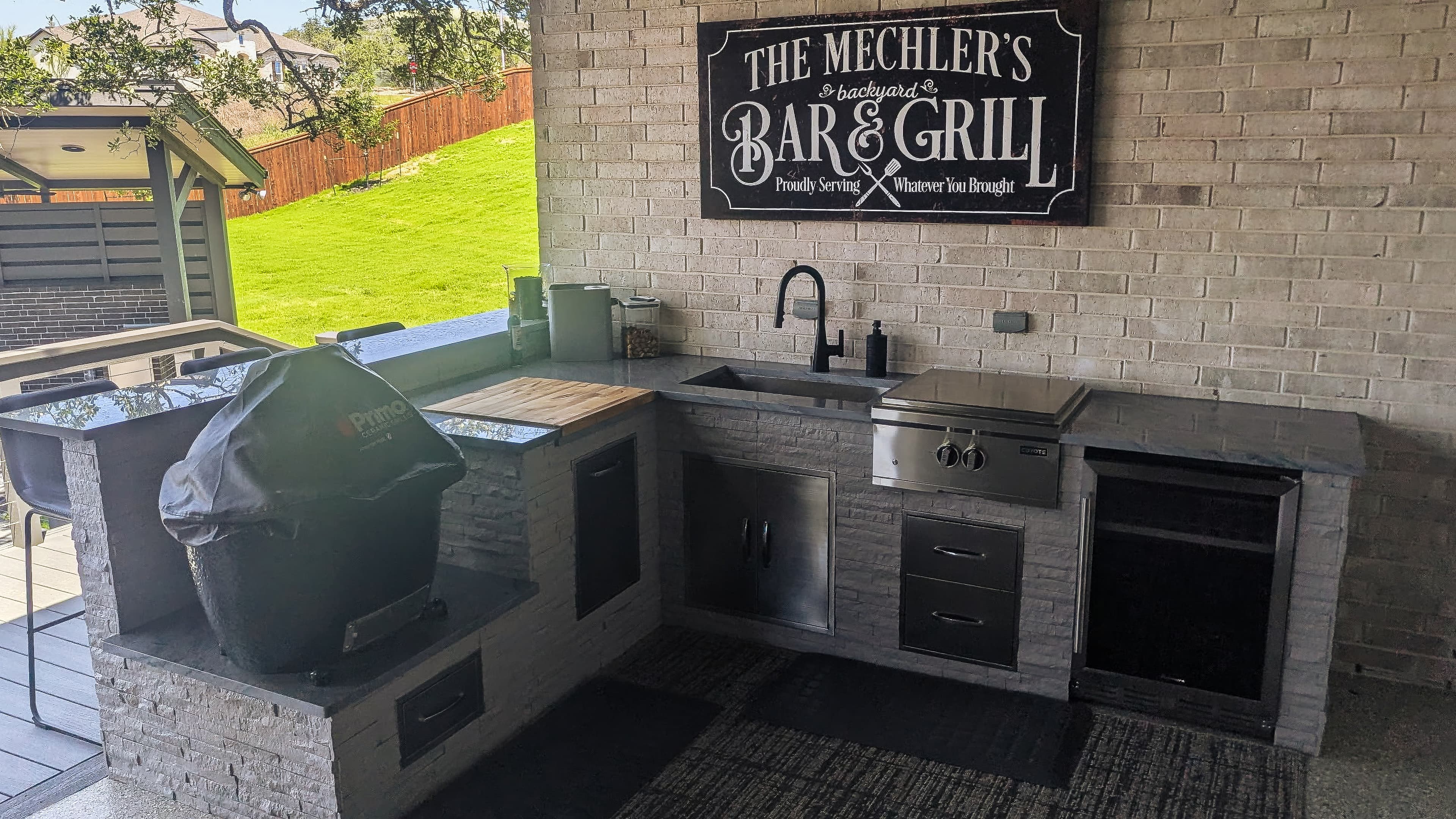 Stone outdoor kitchen with a sink, grill, and personalized backyard bar and grill sign.