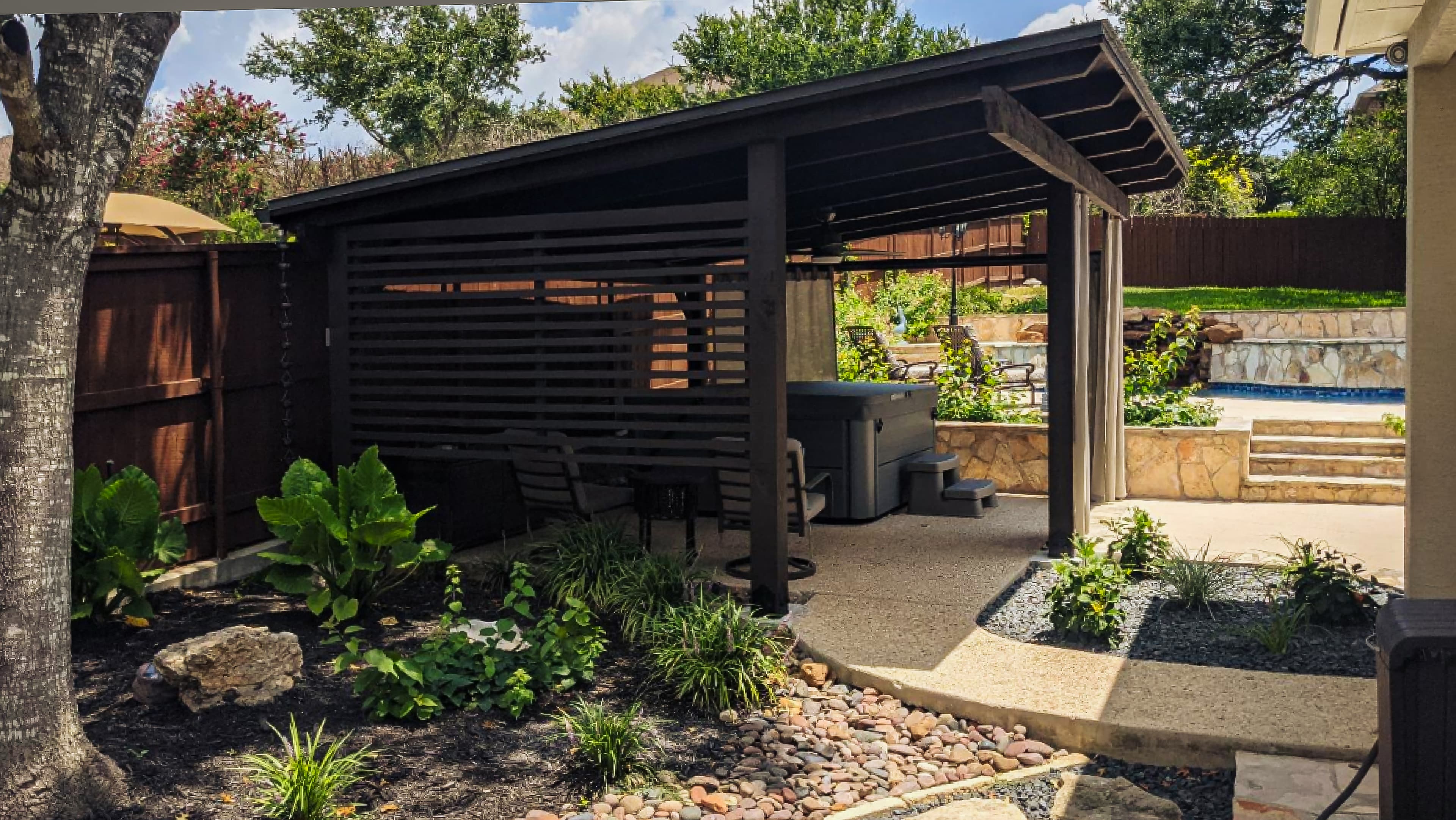 Dark wooden pavilion with a slanted roof sheltering a hot tub in a landscaped backyard.