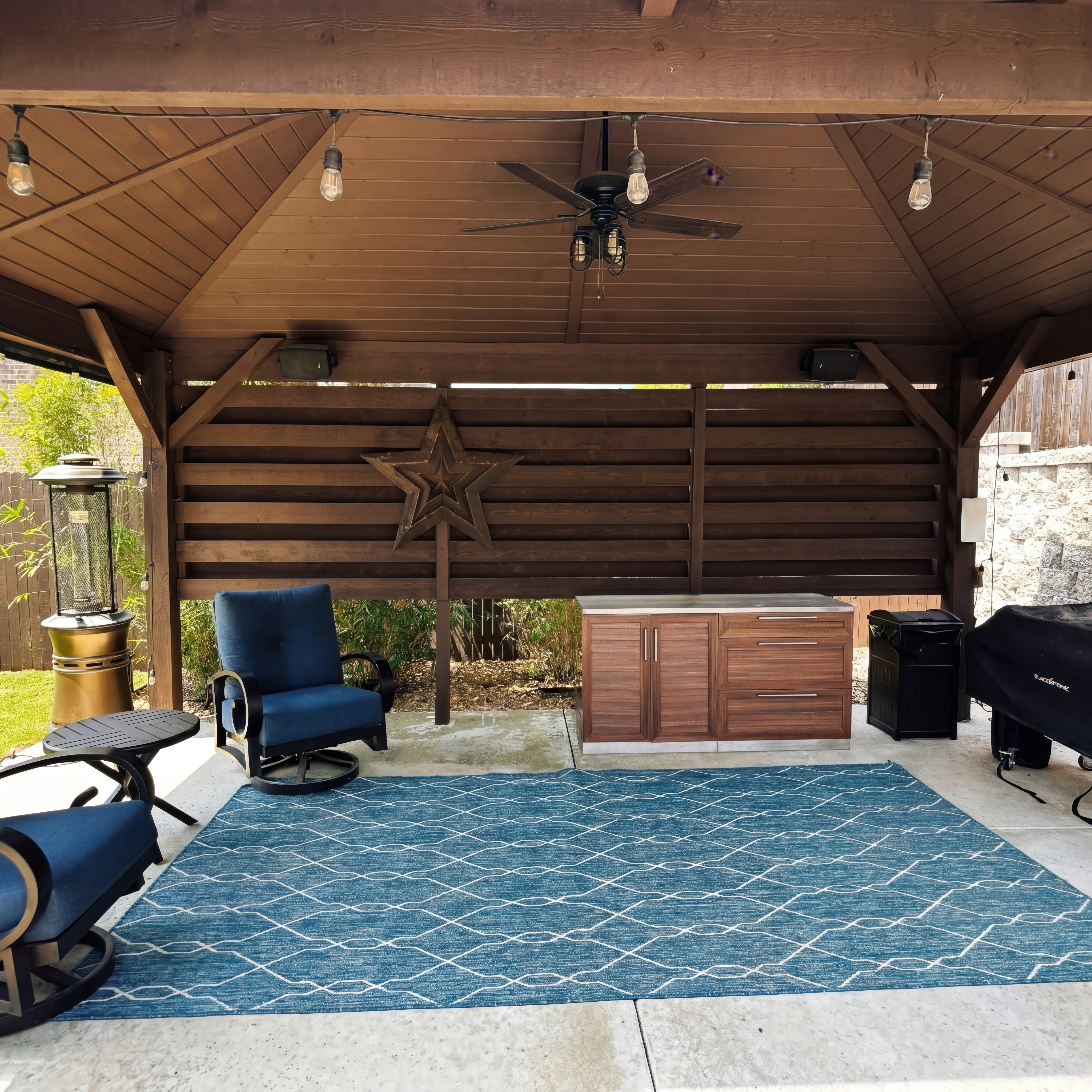 Wooden gazebo featuring blue armchairs, a patterned rug, outdoor kitchen, and decorative wall star.