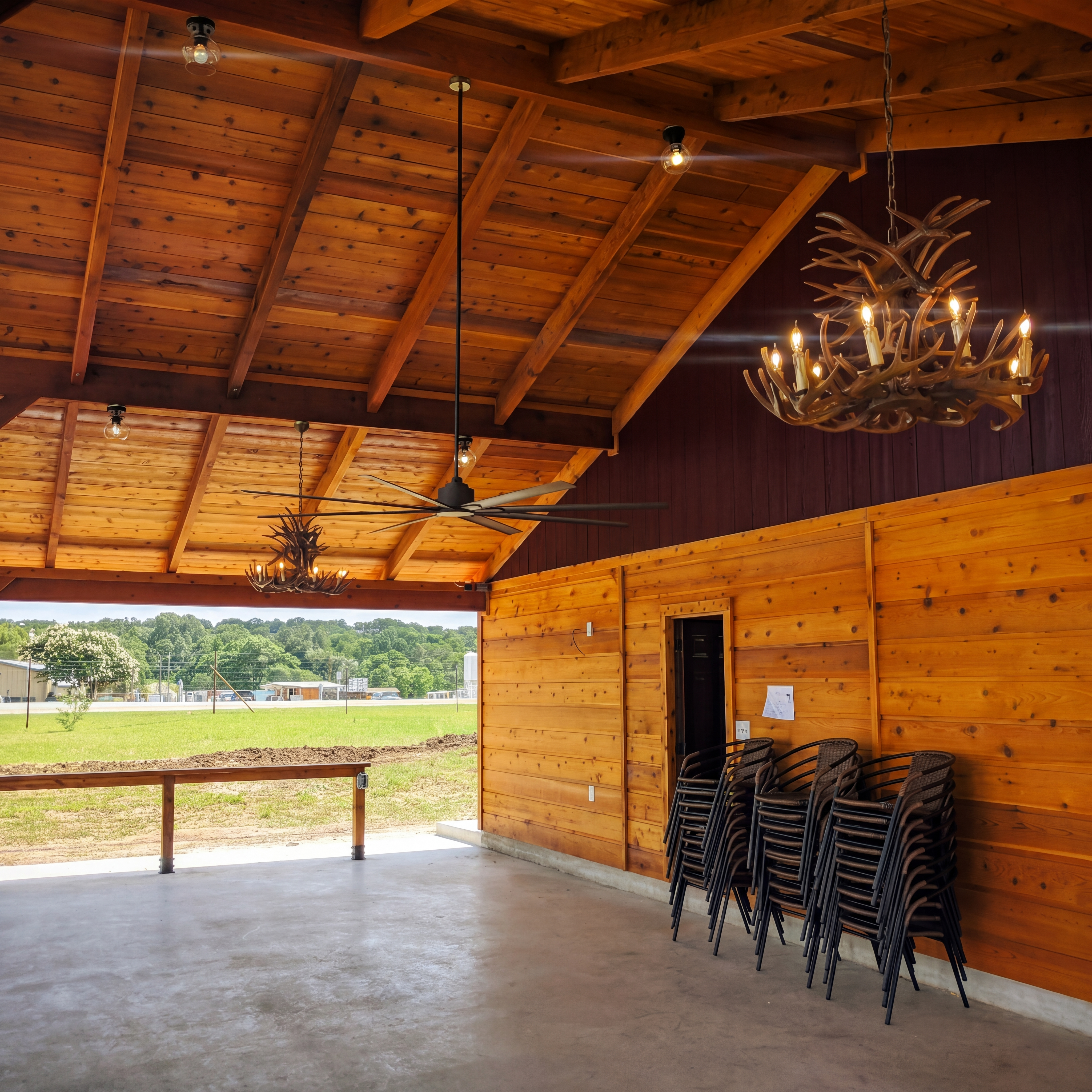 Rustic wooden pavilion with antler chandeliers and stacked chairs overlooking a lush green field.
