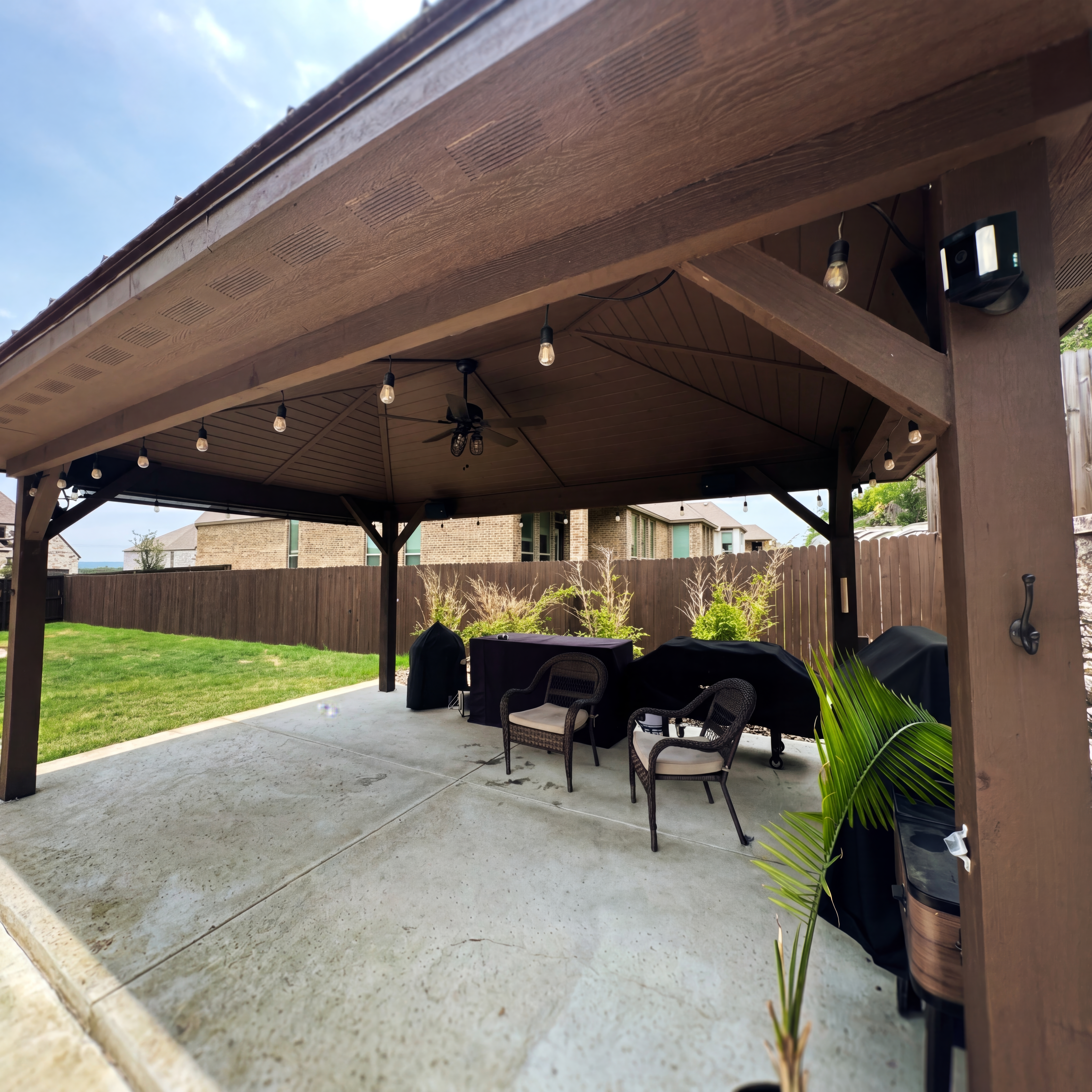 Wooden gazebo with string lights and wicker chairs on a patio overlooking a green lawn.