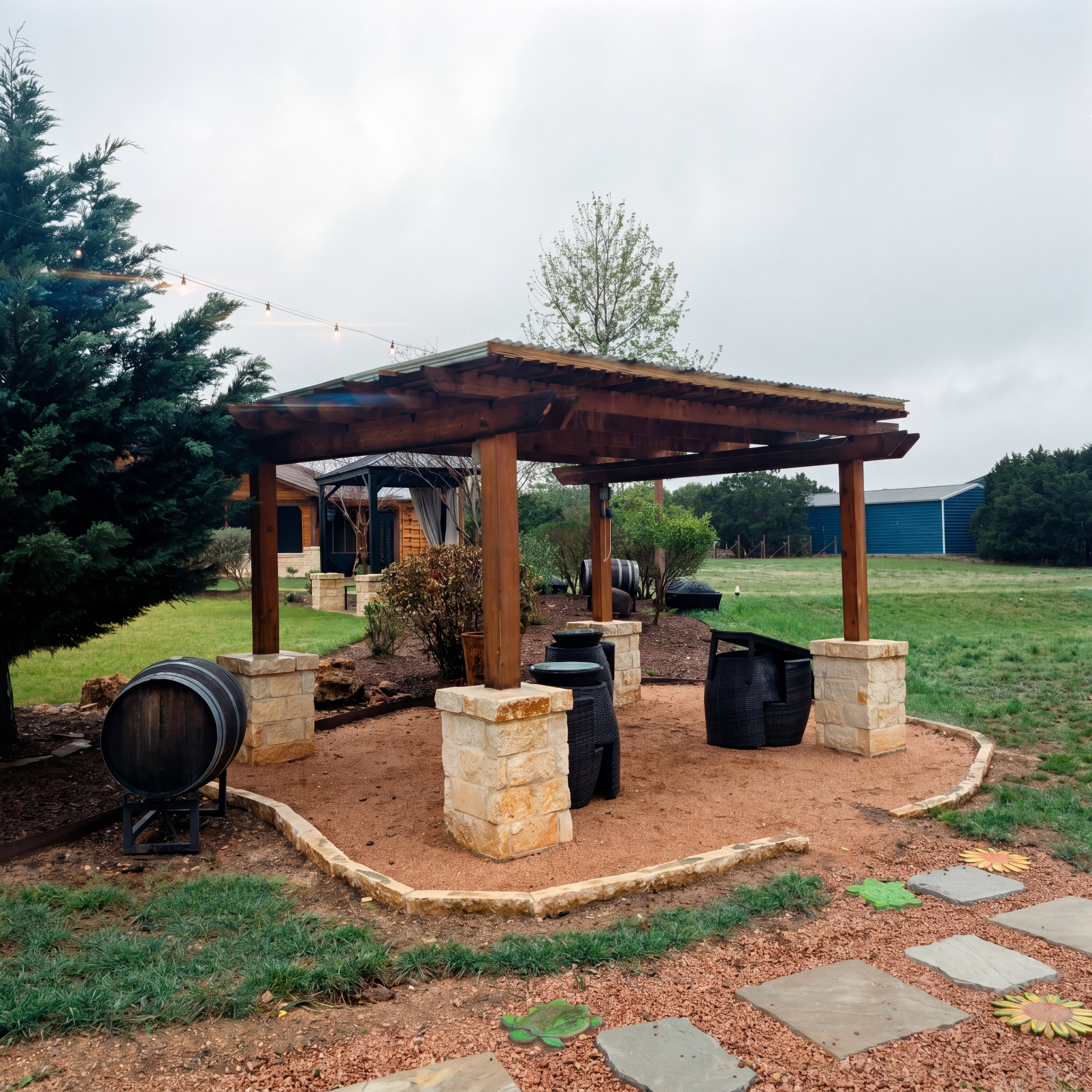 Wooden pergola with stone pillars and black furniture on a gravel patio in a backyard.