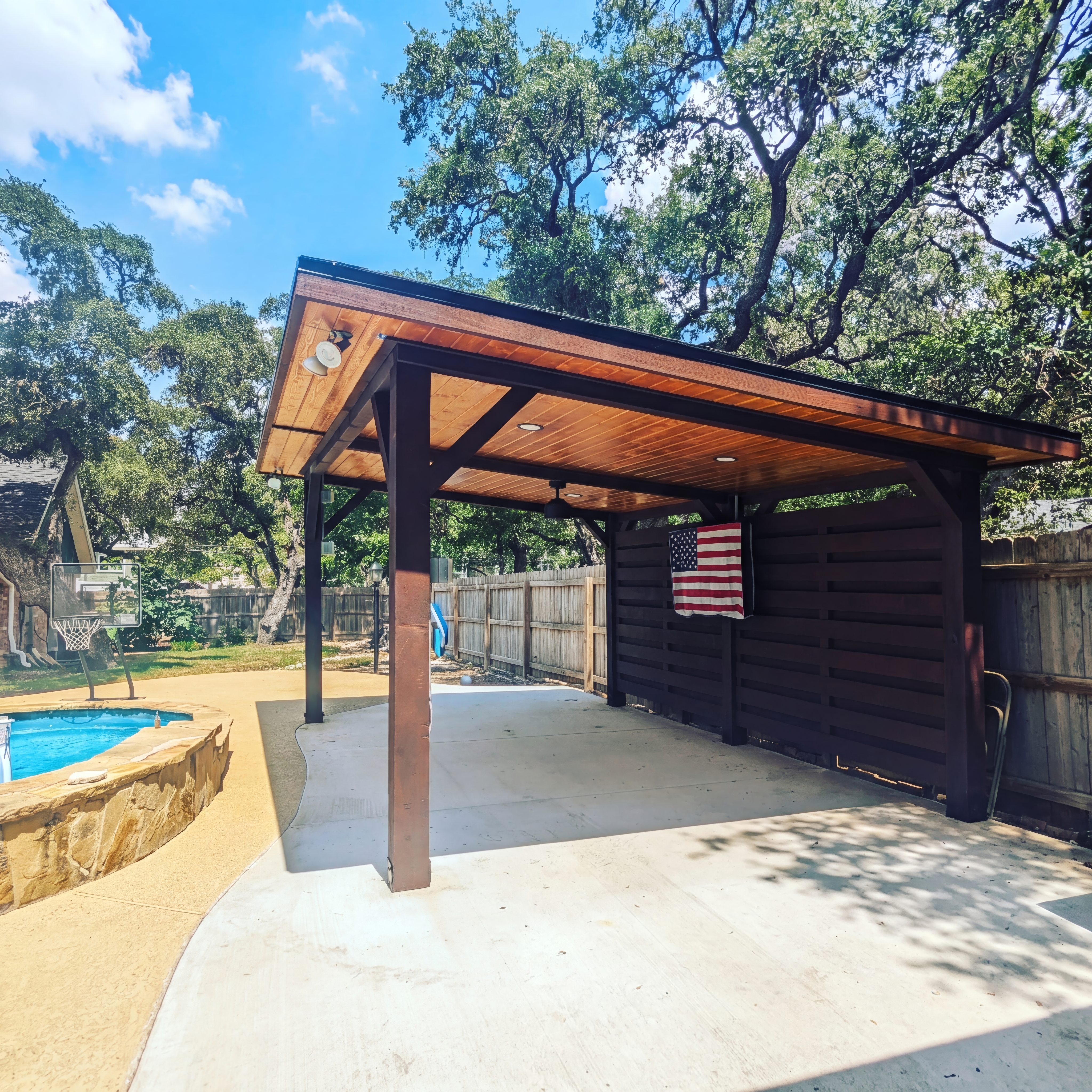 Modern wooden pavilion with an American flag beside a backyard swimming pool.