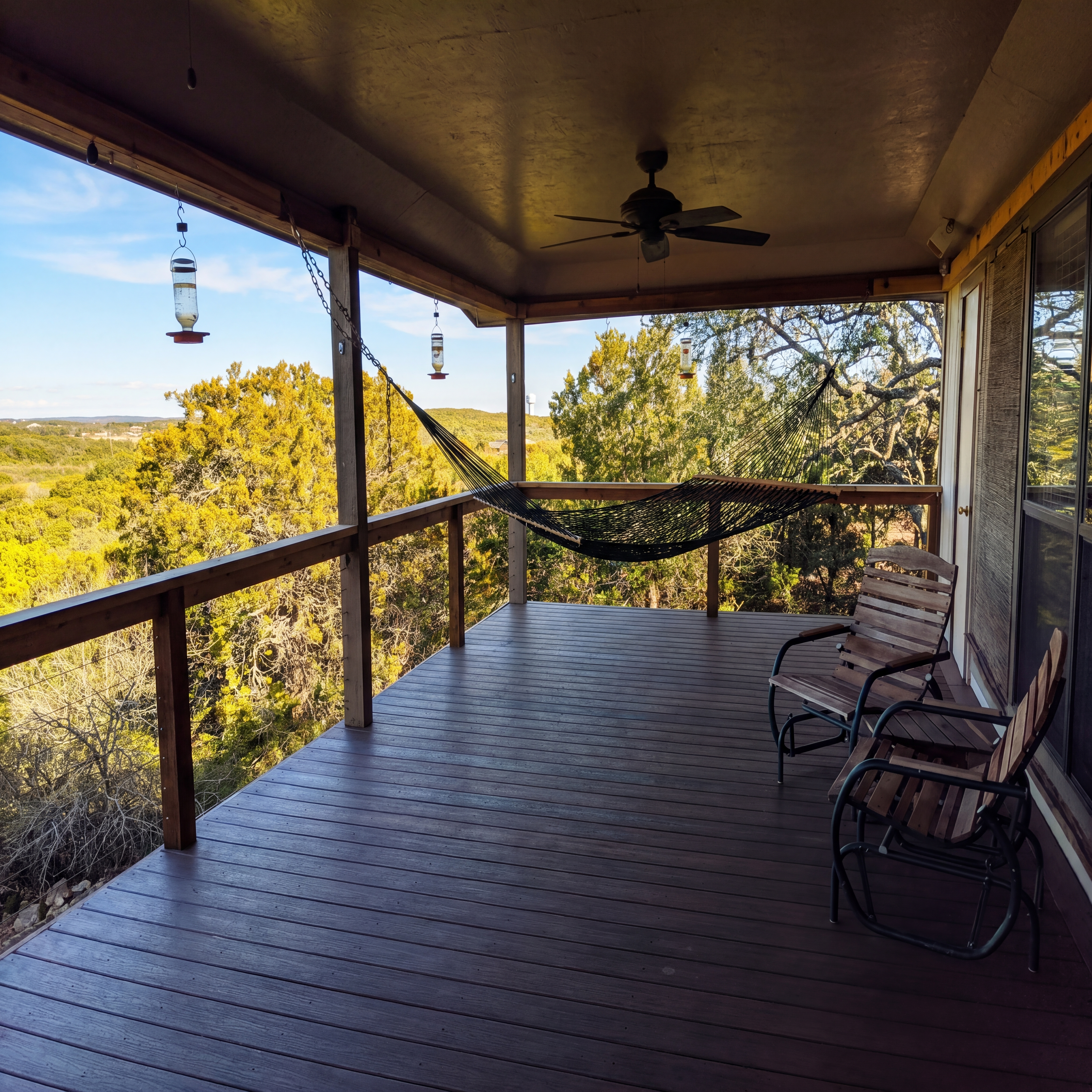 Wooden deck featuring a black hammock and rocking chairs overlooking a lush green forest.