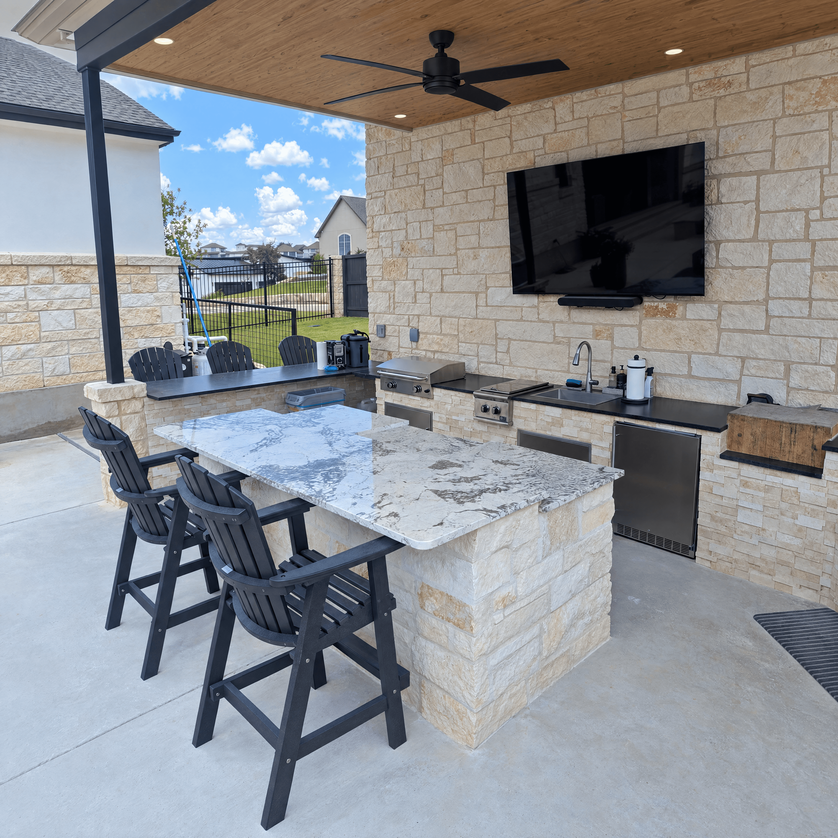 Covered outdoor kitchen with stone island, marble countertop, black bar stools, and wall-mounted television.