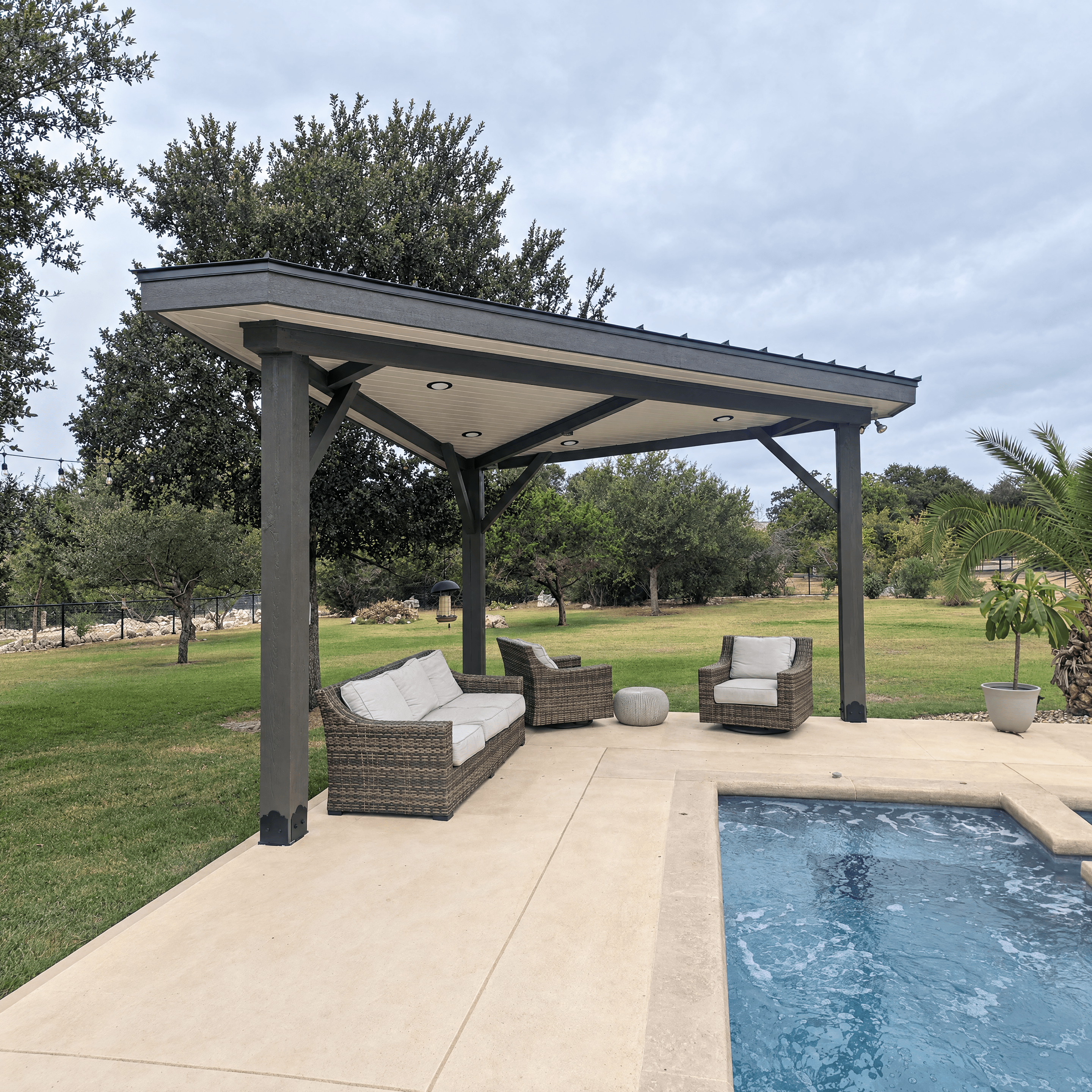 Dark grey pavilion with wicker furniture on a patio beside a backyard swimming pool.